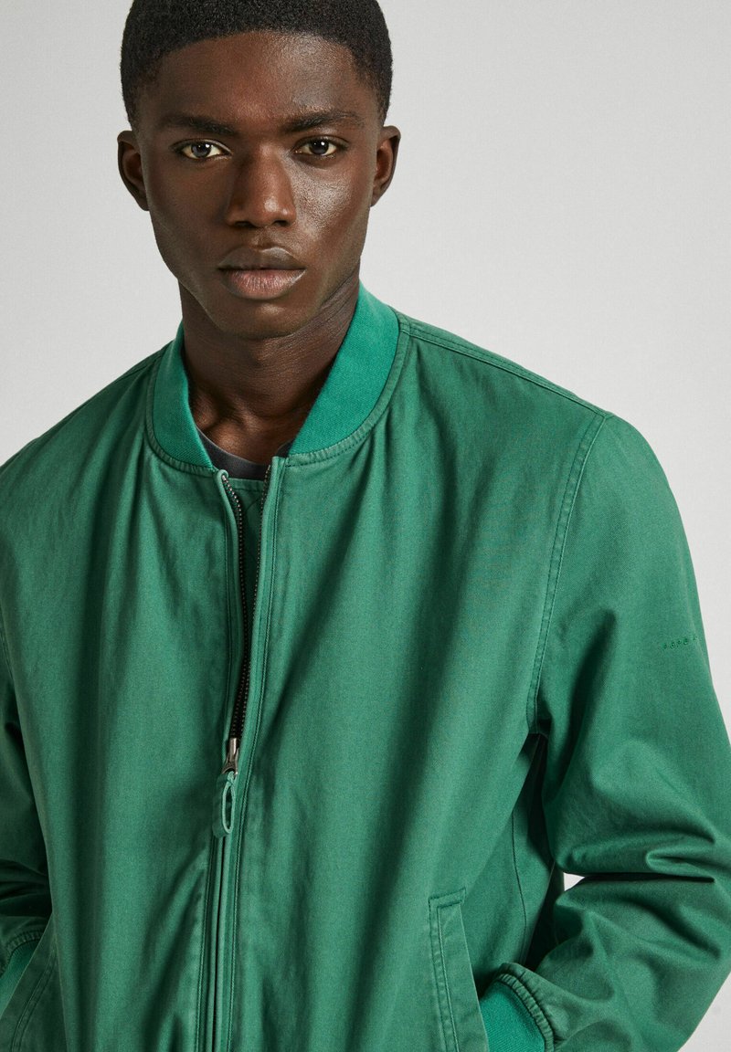 Chaqueta bomber verde con cierre frontal, cuello acanalado y bolsillos laterales. Fabricada en una tela suave con sutiles detalles de costura.