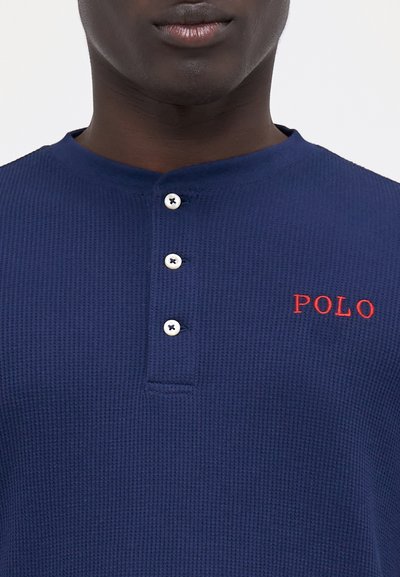 Tums zilā krāsā ar vafelēm teksturētu krekliņu ar četriem pogām, ar vārdiem "POLO" izšūtiem sarkanā krāsā kreisajā pusē. Klasisks apkakles dizains.