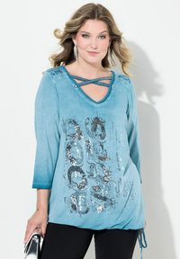 Teal-Oberteil mit langen Ärmeln, V-Ausschnitt und Kreuzdetail. Mit grafischem Druck und Pailletten sowie verstellbarem Kordelzug am Saum.