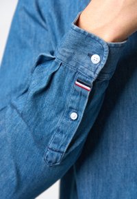 Denim overhemd met een lichtblauw tint. Heeft een met knopen gesloten manchette met twee witte knopen en een klein accent met rood, wit en blauwe strepen.