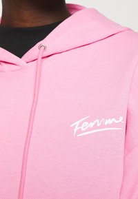 Felpa rosa con un tessuto strutturato, dotata di cappuccio con cordini e di un logo bianco "Femme" in un font stilizzato sul petto.