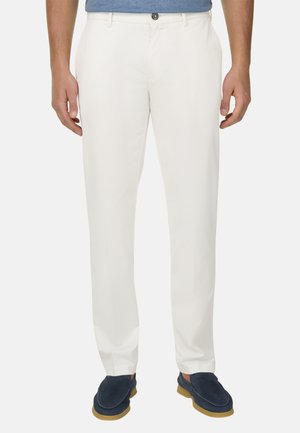 Homme portant un pantalon blanc slim avec des poches avant et des chaussures à enfiler bleu marine sur un fond clair.