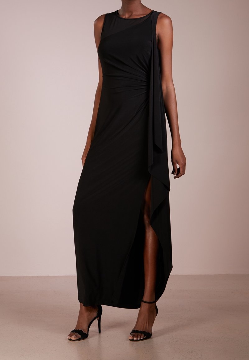 Svart ärmlös maxiklänning med draperad design och sidoklyfta, gjord av lättviktsmaterial. Bärs med svarta högklackade sandaler.