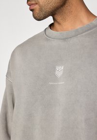 Γκρι φούτερ από απαλό ύφασμα, με λαιμόκοψη crew, διακριτική υφή και λευκό λογότυπο με τα κείμενα "USA" και "FOREVER FORWARD".