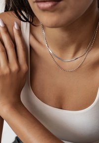 Colliers en argent superposés avec une chaîne serpent plate et une chaîne de perles ronde, présentés sur une peau nue et un débardeur blanc.