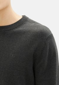 Donkergrijze trui met een ronde hals, fijne breistekstuur en een klein, tonale logo op de borst. Close-up van het schoudergebied.