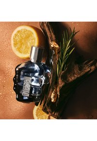 Blå glasflaska formad som en knytnäve, med ett metallock. Omgiven av citronskivor, mörkt trä och grön rosmarin på en strukturerad brun yta.