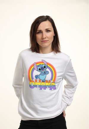Frau mit braunen Haaren trägt ein weißes Sweatshirt, auf dem eine lächelnde blaue Cartoon-Figur mit großen Ohren und der Schriftzug "OHANA" unter einem Regenbogen abgebildet ist.