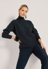 Frau in einem schwarzen gerippten Rollkragenpullover, schwarzen Hosen, mit einem schwarzen Armband und hängenden schwarzen Ohrringen, die vor einer hellen Wand posiert.