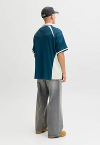 Homme portant une chemise à manches courtes bleu sarcelle et blanche, un jean large gris, des bottes marron clair et une casquette noire, debout, de dos, sur un fond blanc.