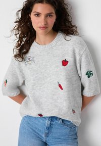 Donna con capelli ricci che indossa un maglione grigio chiaro ricamato con vari ortaggi colorati e jeans blu, in piedi contro un muro bianco.