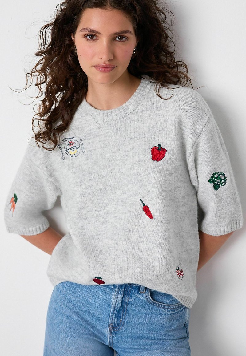 Donna con capelli ricci che indossa un maglione grigio chiaro ricamato con vari ortaggi colorati e jeans blu, in piedi contro un muro bianco.