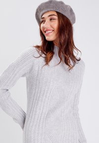 Suéter de punto gris con patrones de trenzas, cuello alto y mangas largas, combinado con una boina de fieltro gris. Textura suave, corte relajado.