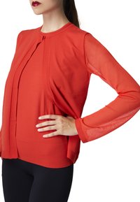 Maglione rosso con maniche lunghe trasparenti, scollatura rotonda e vestibilità comoda, caratterizzato da un dettaglio plissettato sul davanti e un orlo leggermente curvo.
