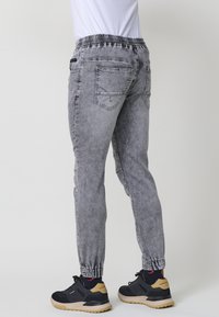 Pantalones joggers de mezclilla gris con cintura y puños elásticos, que cuentan con bolsillos traseros y una textura ligeramente desbotonada. Usados con zapatillas negras.