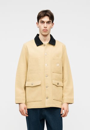 Mladý muž v béžové bundě na knoflíky s černým límcem a velkými předními kapsami, stojící před jednoduchým světlým pozadím.