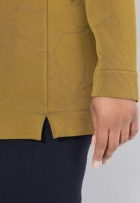 Mano derecha junto a la cintura, llevando una blusa de manga larga mostaza con un sutil patrón floral y pantalones azul marino, contra un fondo gris claro.