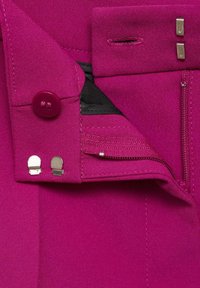 Fuchsia tyg med en slät textur har en knapp och två metallspännen, samt en dragkedjeficka och en sydd söm.