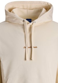 Cremefarvet hoodie lavet af glat stof, med en frontlomme, snøre i hætten og brun broderet tekst på brystet.