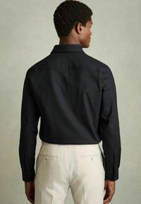 Cămașă neagră cu nasturi, mâneci lungi, cu guler clasic și țesătură texturată. Asortată cu pantaloni bej deschis.