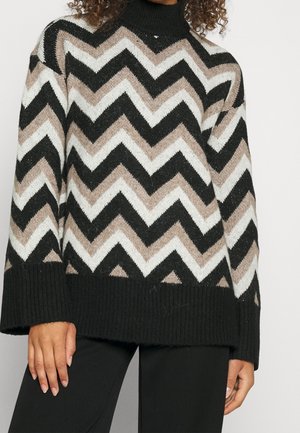 Suéter de cuello alto con patrón de espiga en negro, beige y blanco, con puños y dobladillo acanalados, combinado con pantalones negros.