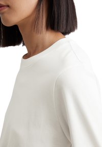 Hvid crew-neck sweatshirt med en glat tekstur, der har en afslappet pasform og skuldersøm detalje, set fra en sideløsning.