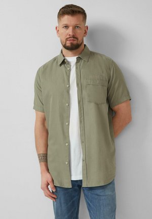 Mann mit Bart, der ein offenes olivgrünes kurzärmeliges Hemd über einem weißen T-Shirt und blauen Jeans trägt, steht vor einer hellen Wand.