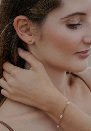 Arany virágmintás tömör fülbevalók, arany láncos és gyöngyös karkötővel viselve, semleges háttér előtt.