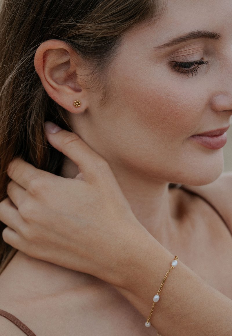 Zlaté náušnice na cvok s kvetinovým dizajnom, nosené spolu s náramkom s pozláteným reťazcom a perlovými akcentmi, na neutrálnom pozadí.