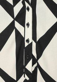 Camicia bianca e nera con un motivo geometrico, dotata di bottoni neri lungo la parte anteriore e cuciture bianche lungo i bordi.