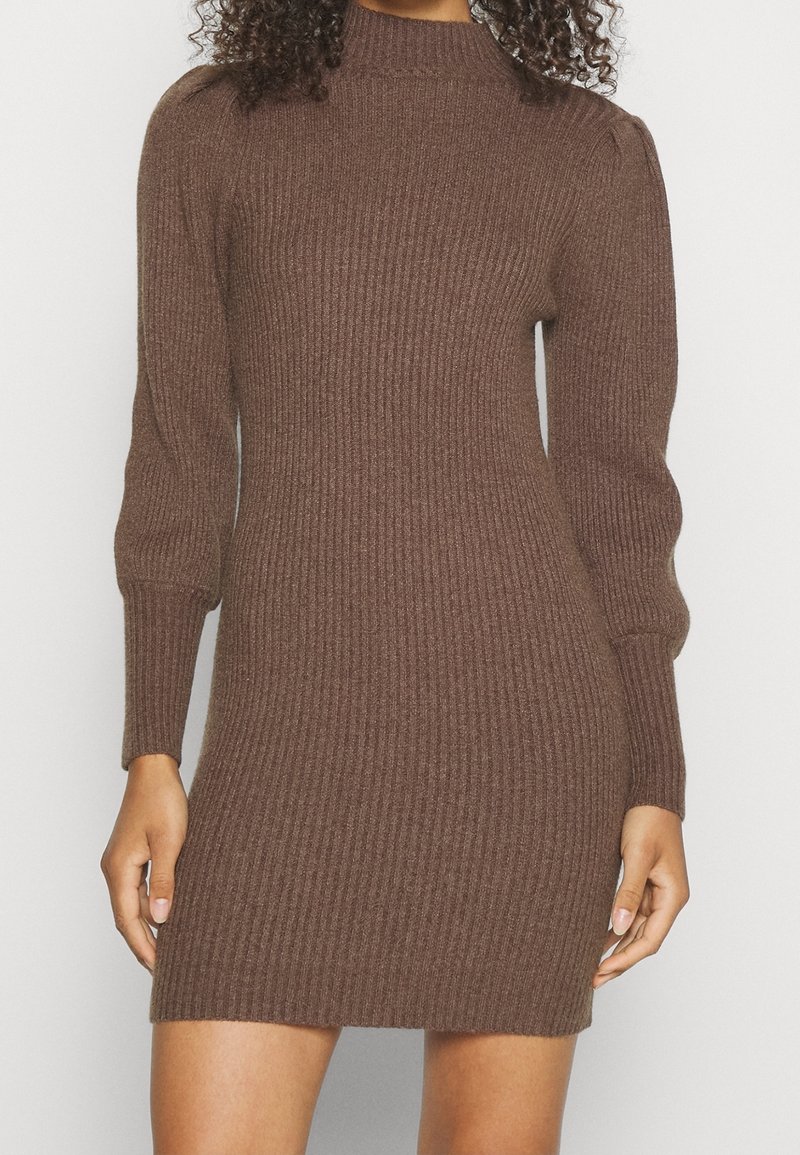 Femme portant une robe-pull en tricot côtelé marron avec des manches longues bouffantes et un col montant, vue du cou jusqu'à mi-cuisse.