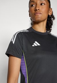 Μαύρο αθλητικό μπλουζάκι με κοντά μανίκια; διαθέτει λευκές ρίγες στους ώμους και μωβ πάνελ στο πλάι με υφή; κατασκευασμένο από λείο ύφασμα.