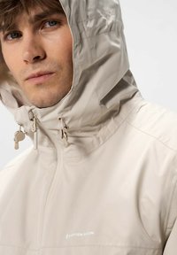 Beige vedenkestävä takki, jossa on huppu, vetoketjusuljenta ja säädettävät narut. Suora pinta ja hillitty brändäys edessä.