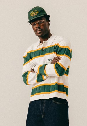 Homme portant des lunettes, une casquette verte et un pull rayé blanc, vert et jaune, debout les bras croisés devant un fond uni.
