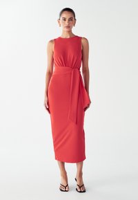 Figurbetontes, ärmelloses rotes Midikleid mit Taillengürtel, getragen mit schwarzen offenen Absatzsandalen, abgebildet auf einer Frau vor einem schlichten Hintergrund.