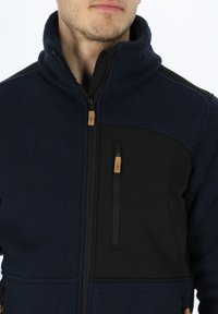 Giacca in pile di un blu navy scuro con dettagli neri. Presenta un colletto alto, tasche con zip e un tessuto testurizzato. Design resistente per l'uso all'aperto.