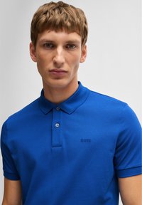 Plava polo majica od teksturirane tkanine, s dvostrukim gumbom i suptilnim "BOSS" logom na lijevoj strani prsiju. Kratki rukavi.