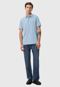 Polo azzurro con tre bottoni, maniche corte e un logo discreto. Abbinato a jeans dritti blu scuro e scarpe nere.