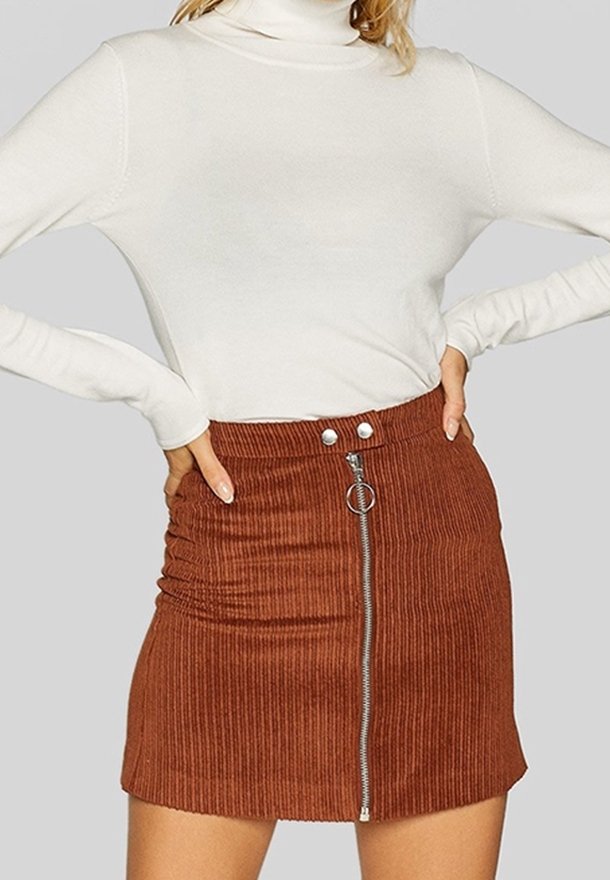 Mini-jupe en velours côtelé marron avec fermeture éclair à l'avant, deux boutons supérieurs et texture froncée sur les côtés. Associée à un haut blanc à manches longues.