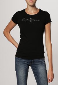 Nainen, jolla on yllään tiukka musta lyhythihainen t-paita, jossa on kimaltelevaa tekstiä, ja siniset farkut. Hän seisoo käsi lantiolla.