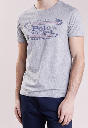 Mand iført grå Polo Ralph Lauren T-shirt med mørkeblå tekst og mørkeblå bukser, stående mod en lys baggrund.