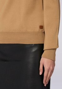 Maglione beige lavorato a maglia con polsini a coste, dotato di un'etichetta in pelle marrone. Abbinato a una gonna in pelle nera, che mostra una superficie liscia e un design aderente.