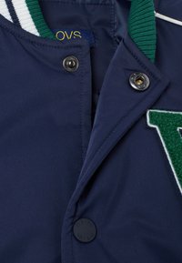 Giacca blu navy con colletto a costine verde, dettaglio in feltro verde con lettere prominenti e bottoni a pressione. Presenta un'etichetta logo all'interno.