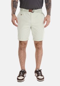 Pantaloni corti verde chiaro con risvolti, una cintura intrecciata e tasche frontali. Abbinati a sneakers bianche e marroni, che mostrano uno stile casual.