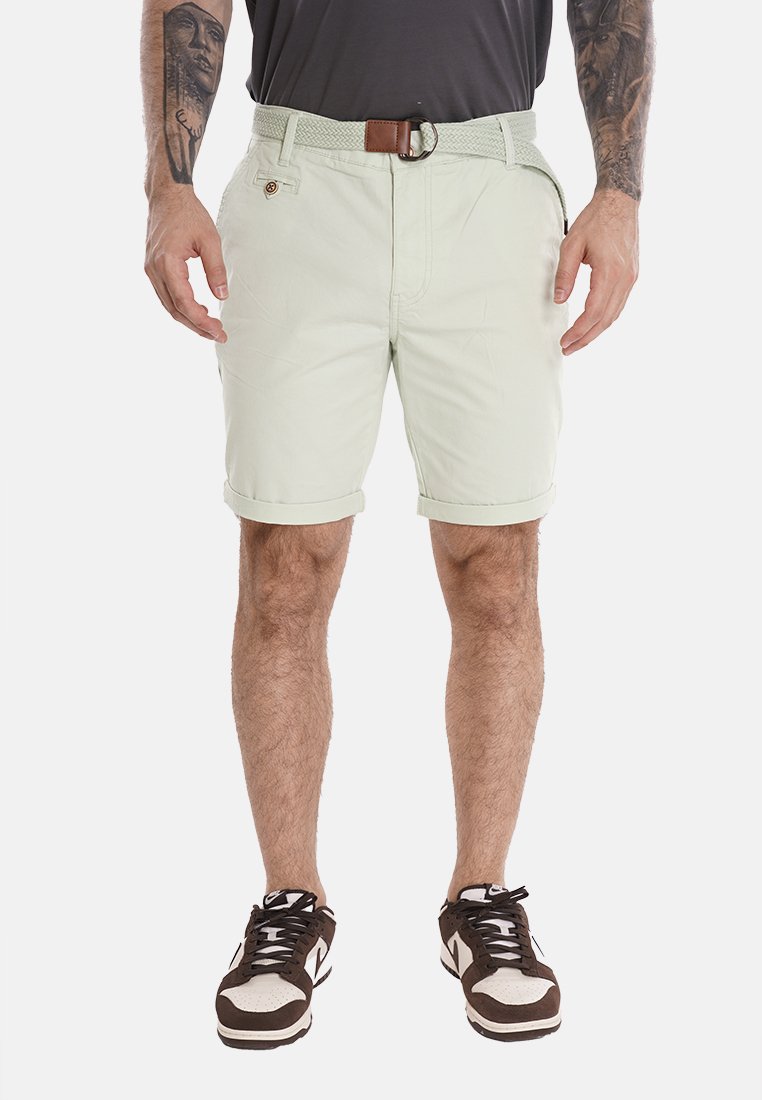Pantaloni corti verde chiaro con risvolti, una cintura intrecciata e tasche frontali. Abbinati a sneakers bianche e marroni, che mostrano uno stile casual.