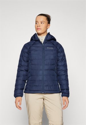 Giacca puffer blu navy con cappuccio, con motivo trapuntato, cerniera frontale e logo sul lato sinistro del petto; abbinata a pantaloni beige.
