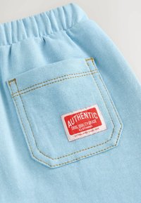 Shorts en tissu bleu clair avec une taille élastique, dotés d'une poche arrière avec une étiquette "AUTHENTIC" rouge et blanche, et des accents de couture jaunes.