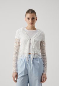 White lace cropped cardigan with long sleeves, featuring a hood and tied closure at the front. Worn over a white top and paired with light blue pants.