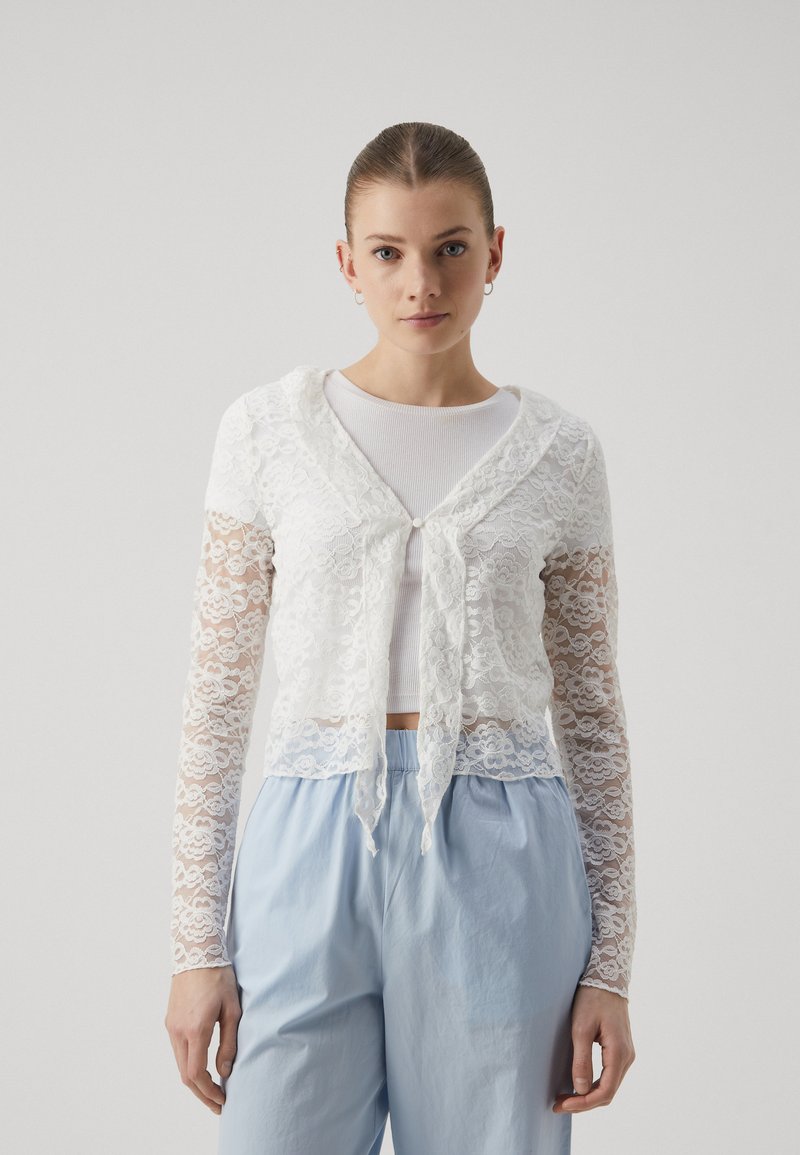 White lace cropped cardigan with long sleeves, featuring a hood and tied closure at the front. Worn over a white top and paired with light blue pants.