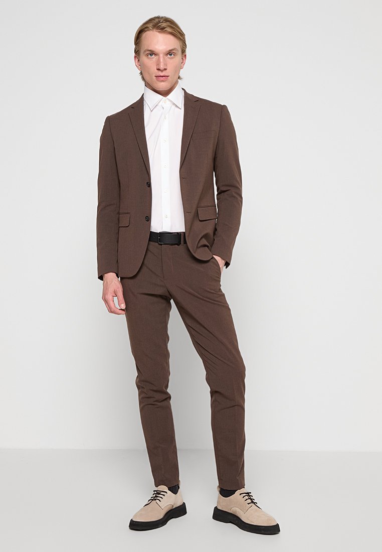 Un jeune homme se tient contre un mur blanc, vêtu d'un costume marron, d'une chemise habillée blanche, d'une ceinture noire et de chaussures beiges avec des semelles et des lacets noirs.